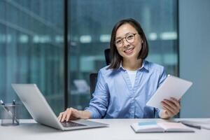 Portrait of young Asian businesswoman, office worker smiling and looking at camera, working remotely with colleagues, holding tablet computer in hands. photo