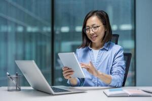 Asian woman dressed professionally, using a tablet at her desk in office. She appears focused and engaged, demonstrating concepts of technology usage and productivity in professional environments. photo