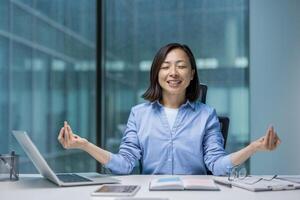 Young joyful Asian woman meditating, resting and relaxing in lotus position. Woman smiling contentedly doing breathing exercises. Businesswoman inside office at workplace, calm and focused. photo