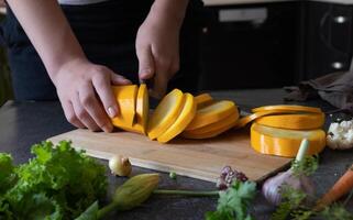Hands are shown slicing a yellow squash on a cutting board, amidst an array of fresh vegetables in a kitchen setting photo