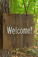 A rustic wooden welcome sign hanging from a tree in a forest photo