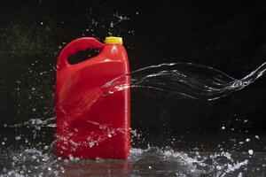 Front view of red plastic motor oil bottle splashed by stream of water, container with handle and yellow screw cap without label photo