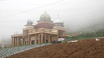 A mosque with a large dome stands in the middle of hills covered in thick fog. This mosque has a unique architecture with small towers on each corner. Surrounding it are agricultural fields. photo