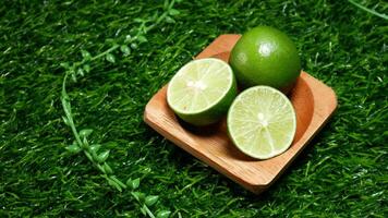Fresh limes, consisting of one whole lime and two slices, neatly arranged on a small wooden plate. With synthetic green base grass and decorative leaves enhance the natural. photo
