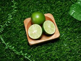The lime consists of one whole lime and two slices, neatly arranged on a small wooden plate. With a base of synthetic green grass and ornamental leaves that complete the natural impression. photo