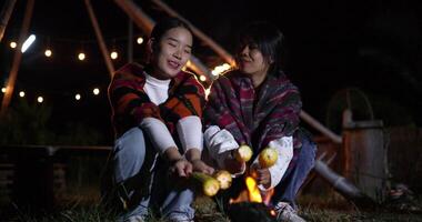 Handheld low angle view shot, Two Asian siblings enjoy talking and grilling corn while sitting around a warm campfire on a chilly evening. video