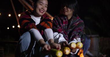 Handheld selective focus shot, Two Asian siblings enjoy talking and grill corn while sitting around a warm campfire on a chilly evening video
