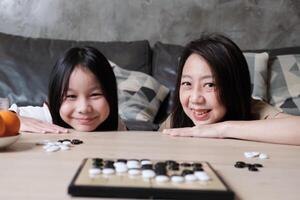 Portrait of Asian family, mum, and kid fun playing Go, checkers board game on table in home living room with, happy weekend times relaxing hobby, smile and look at camera together. photo