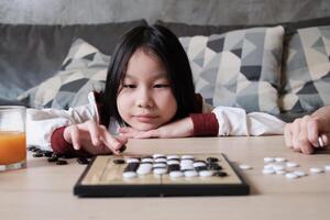 One Asian child fun playing Go, checkers board game on table in home living room, cheerful smile and looking at camera, happy weekend relaxing hobby of clever cute child winner. photo