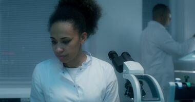 A female laboratory assistant looks up from her work and looks at the camera with a serious look, sitting in front of a microscope in a medical laboratory video