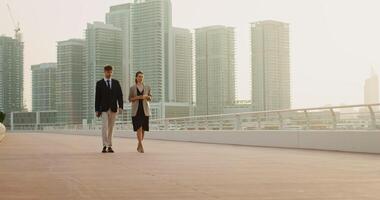 A man with glasses with a folder in his hands and a woman with a disposable cup, dressed in business clothes, walk along the embankment and talk with a serious look video