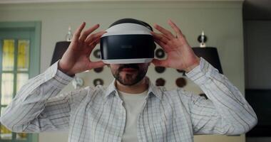 A man in casual clothes puts on a virtual reality headset while looking at the camera, and starts an online game while standing at home. video