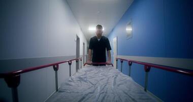 An orderly carries an empty gurney down the hospital corridor walking backwards video