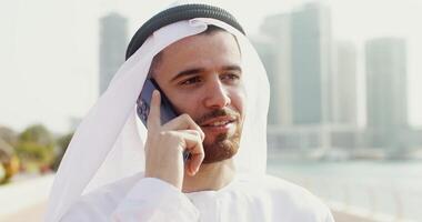 A young man in Arab national white clothes smiles while talking on a mobile phone while standing in the open air. Face close-up video