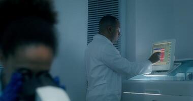 A man in a lab coat sets up a blood splitting machine. A woman uses a microscope sitting at a table. Working in a medical laboratory video