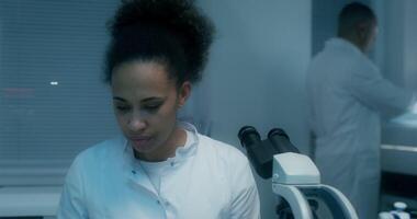 A female laboratory assistant looks up from her work and smiles at the camera, sitting in front of a microscope in a medical laboratory video