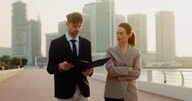 A man and a woman in business clothes study documents lying in a folder walking along the embankment in a modern metropolis video