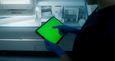 A man in a lab coat and disposable gloves uses a tablet with a green screen standing in a medical laboratory, close-up of his hands, an unrecognizable person video