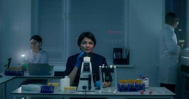 A woman in a lab coat smiles at the camera sitting in front of a microscope in the laboratory, working next to her colleagues video