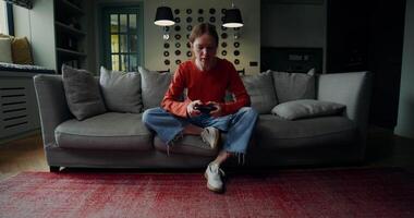 A young woman in casual clothes is smiling while playing games with a joystick, sitting on the couch at home. The moves from the general plan to the woman's face video