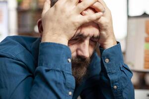 Closeup of male analyst holding his head in hands, having exhausted expression from reviewing financial analysis. Bearded businessman with eyes closed, feeling tired and overwhelmed in startup office. photo