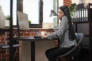 Female manager multitasks during phone call with client, entering details on computer in startup office. Asian woman seated at her desk, using keyboard and speaking with colleague on mobile device. photo