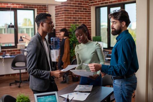 Coworkers standing around table and discussing strategies for company growth in startup office. Multiethnic business people at desk with papers in their hands, reviewing and comparing research data. photo
