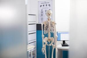 Human skeleton stands ready for diagnostics in unoccupied hospital room. Skeletal structure placed in doctors office with computer, medical bed and cabinet suggest a focus on orthopedic care. photo