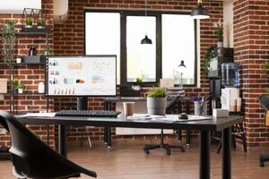 Table in spacious startup office holds desktop pc showing financial analytics. Brick wall interior includes digital devices, shelves, water cooler, and plant decorations, creating refreshing workspace photo