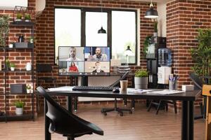 Empty brick wall office has desktop screen showing colleagues on videoconference. Well arranged room containing shelves, indoor plants, water dispenser and computer displaying ongoing virtual meeting. photo