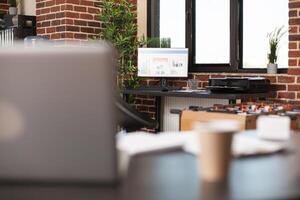 Brick wall room with financial charts on computer, printer on desk surrounded by neat furniture and plant decoration. Startup office set with laptop, plants and desktop pc displaying business data. photo