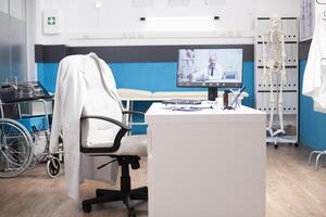 Empty hospital office setup with desktop monitor displaying an online medical conference. Computer screen shows caucasian doctor offering remote healthcare consultation in well-arranged clinic room. photo