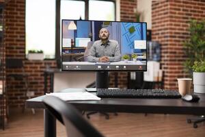 Caucasian man videocall displayed on desktop monitor positioned on table of vacant brick wall workspace. Office desk holding computer with active virtual session showing businessman doing presentation. photo