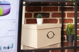 Closeup of box placed on shelf with plant decorations and digital screen nearby. Selective focus on office container kept on cabinet as device displays marketing data in modern brick wall room. photo