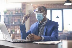 African american man seated at desk, focused on startup analytics with pen in hand. Black male manager in brick wall office tapping his forehead and strategizing new business projects on laptop. photo