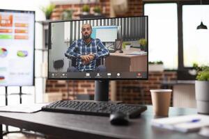 Vacant table with desktop screen displaying online session with businessman in wheelchair in spacious, arranged office setup. Brick wall room is equipped with computers and business planning supplies. photo