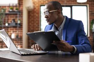 Businessman with spectacles is holding company research papers and checking data on laptop. Male analyst seated at office desk updating business analytics on device with information from clipboard. photo