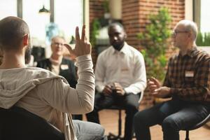 Selective focus on young guy making gestures with his hand during therapy session. Group of diverse individuals is seated in brick wall room, discussing mental health struggles and progress. photo