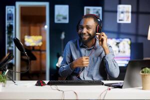 Cheerful BIPOC internet show host in studio uses camera to film wired headphones review, happy with crisp sound. Influencer satisfied by noise cancelling effect on stereo music listening device photo