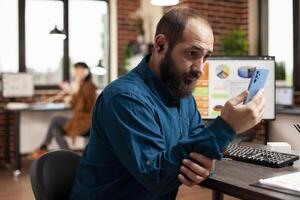 Male employee using smartphone to check social media as his desktop monitor shows market analysis. Closeup of bearded man with earphones, sitting in startup office and holding his mobile device. photo