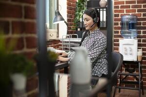 Female entrepreneur using headphones types on her desktop pc in an organized startup office. Asian woman advisor with a wired headset working on her computer and speaking to clients. photo