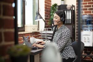 Female employee uses desktop pc, typing on keyboard and engaging in a lively conversation through wired headphones in modern workplace. Smiling woman seated at office desk, using computer and headset. photo