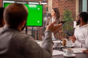 Diverse team looking at greenscreen display on digital device. Caucasian woman with clipboard standing near monitor with isolated mockup template at business meeting with colleagues in office. photo