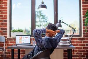 Black entrepreneur stretches his back in modern startup office, feeling accomplished after completing tasks. Male analyst with face mask, leaning in his chair and reflecting on productive day at work. photo
