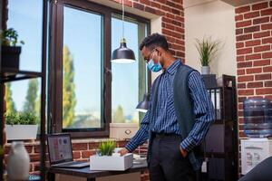 Male employee adjusts plant and work materials in box and reviewing financial documents on table near window in his workspace. African american manager finalizing his tasks before leaving the office. photo