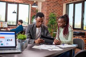 Businessman explaining marketing strategy on tablet to female coworker in brick wall workspace. African american colleagues sitting at table with documents and having a serious conversation in office. photo