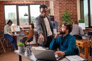 Male analyst points at laptop screen, explaining market analysis update to caucasian coworker. Bearded man sits and listens attentively to black manager describe financial errors in his presentation. photo
