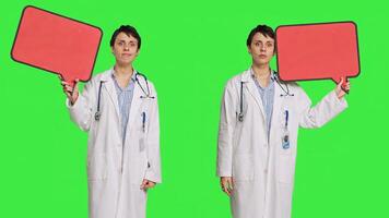 Woman doctor holding a red empty speech bubble sign in studio, showing an isolated blank cardboard icon against greenscreen backdrop. Physician creating announcement with banner. Camera B. photo