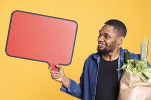 African american guy holds a speech bubble empty icon to create an ad for zero waste and sustainable lifestyle. Doing a promotional marketing advertisement for fresh bio fruits and veggies. photo