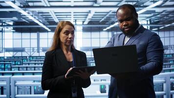 Coworkers doing yearly maintenance using tablet and laptop to prolong data center equipment life span. Team of admins in server hub workplace monitoring supercomputers using digital device, camera B photo
